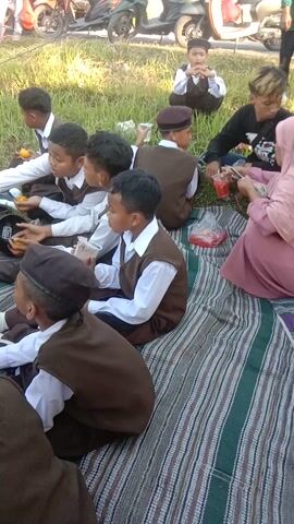 The video shows a group of young children, likely students, wearing brown vests and white shirts, and some wearing brown hats, sitting on a striped mat in an outdoor grassy area. The children appear to be engaged in some activity, possibly a snack break or a lesson. Several children are holding oranges and some are looking down at their hands or at objects on the ground. There are also adults present, one of whom is wearing a light brown hijab and glasses, and another adult in the background wearing a pink garment. Several motorcycles are parked nearby, along with shoes and bags scattered on the ground. The lighting suggests it is daytime, possibly late morning or early afternoon, with sunlight casting shadows. The overall atmosphere appears to be casual and communal.