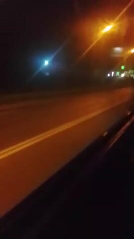 A low-angle, handheld video shows a dark, wet road at night. Bright streetlights cast a yellow and white glow over the scene, creating lens flares and reflections on the wet surface. White lane markings are visible on the road. In the distance, there are some blurred lights and structures, suggesting an urban or roadside environment. The video appears to be shot from a moving vehicle.