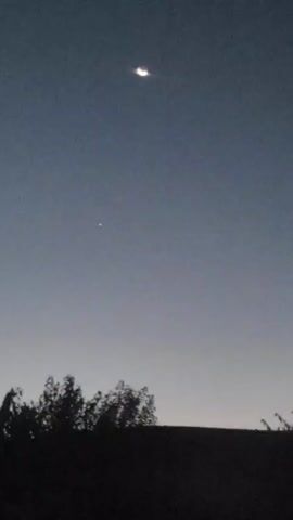 A wide shot shows a night sky with a sliver of a crescent moon high up and a small, bright star or planet beneath it. Silhouetted trees and a dark horizon line are visible at the bottom of the frame. The sky transitions from a deep blue at the top to a lighter, hazy blue near the horizon. There are no people or discernible actions taking place.