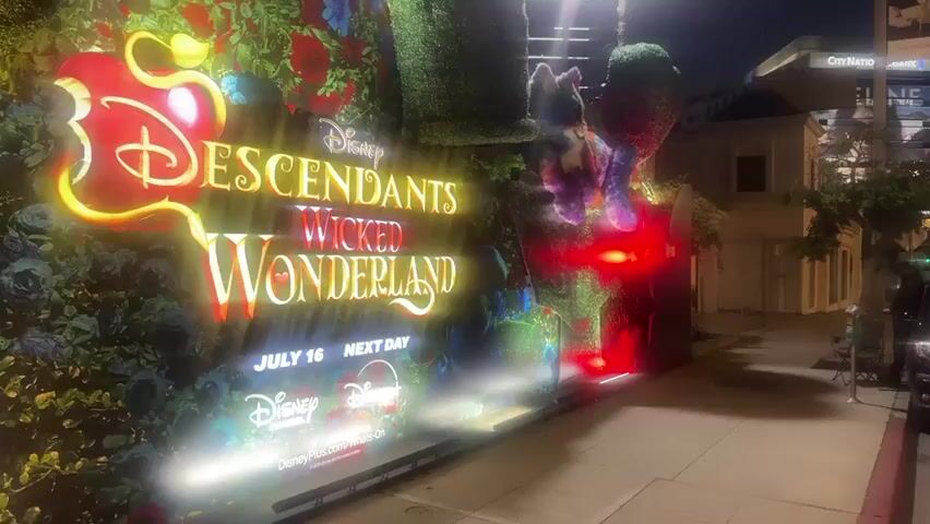 The video shows an outdoor display at night, illuminated by bright lights. The display features a large sign that reads "Disney DESCENDANTS WICKED WONDERLAND" in glowing yellow and orange letters. The sign is surrounded by a backdrop of lush green foliage, red roses, and blue flowers, creating a whimsical and fantastical scene. In the upper right of the display, a fluffy, colorful creature, possibly a cat, is perched on a topiary. To the right of the main sign, there is a stone archway that frames a vibrant, colorful light display of red and green, resembling a stained-glass window. The ground in front of the display is a paved sidewalk, and a street is visible to the right. The overall atmosphere suggests a promotional event or installation for the Disney movie.