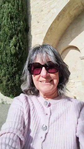A woman wearing sunglasses and a light pink sweater stands in front of an arched stone building. She speaks. A tall green bush is visible to the left. The sun is shining.