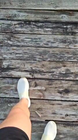 The video begins with a close-up shot of a weathered wooden surface, possibly a dock or pier. The wood planks are worn and gray, with some green patches of moss or grass visible. The camera then pans to reveal a body of water, reflecting the soft colors of the sky. The water is calm, with some lily pads and reeds growing near the edge. In the distance, a treeline is visible against the horizon. The sky transitions from a pale blue to shades of orange and pink as the sun begins to set, casting a warm glow over the scene. A small wooden dock or pier extends into the water, with its reflection clearly visible on the surface. The overall impression is one of tranquility and natural beauty at dusk.