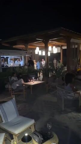 The video captures a lively evening scene at a beachfront establishment, likely a restaurant or bar, at Playa Del Sol-Villacana, Spain. The time of day is clearly night, indicated by the dark sky and the artificial lighting illuminating the area.

The setting is a sandy beach, with seating areas arranged on the sand. In the foreground, several chairs and small tables are visible, some with drinks on them. One chair is particularly prominent, with a striped cushion. In the mid-ground, a covered outdoor seating area with a wooden roof is a central focal point. It is illuminated by several pendant lights, creating a warm and inviting ambiance.

Several groups of people are present, engaged in various activities. Some are seated, relaxing and conversing, while others appear to be standing and mingling. Towards the back of the scene, there's a bar area with people standing behind it, possibly serving drinks. In the covered area, a group of people are gathered, and some appear to be dancing, suggesting a celebratory or social event is underway. The overall mood seems convivial and relaxed, with the warm lighting and the presence of people enjoying themselves contributing to a pleasant atmosphere.

Notable details include the outdoor furniture, which appears to be made of natural materials like bamboo and rattan, fitting the beachside location. The plants in pots scattered around the seating areas add to the natural and tropical feel. The lighting is a significant element, with the pendant lights and smaller table lamps creating intimate zones within the larger space. The presence of wine glasses and a handbag on a table suggests a social gathering or dining experience. No visible text is present in the video.