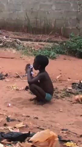 A young, dark-skinned child sits in the dirt in what appears to be a poor outdoor area. The child is shirtless, wearing light green shorts. The child sits squatting, holding what seems to be a small, crushed aluminum can. The ground is reddish-brown dirt littered with trash. A brick wall is in the background, with some grass and weeds growing nearby.