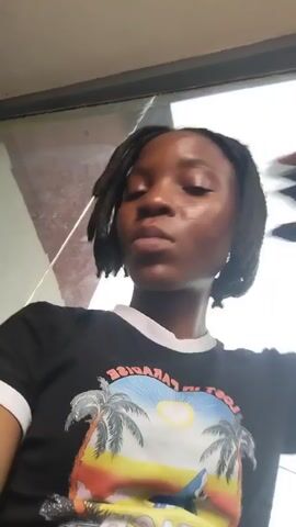 The video shows a close-up, selfie-style shot of a young Black woman with dark braided hair. She is wearing a black t-shirt with a graphic print featuring palm trees, a beach scene, and text. The woman's skin appears shiny, possibly from sweat or makeup. She is looking directly at the camera with a neutral expression. Her right hand is raised and resting on the back of her head. The background is blurred, but it appears to be outdoors with some structures and possibly vehicles visible. The lighting suggests it is daytime. The video consists of a few seconds of footage where the woman's expression and hand position change slightly.