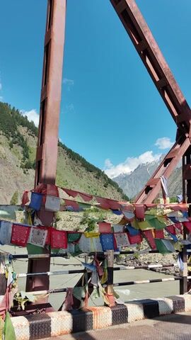 This media file captures a view from a rusty, red metal truss bridge, adorned with colorful Tibetan prayer flags. The flags, in hues of blue, white, red, green, and yellow, are strung along the sides of the bridge, fluttering in the wind. 

The scene is set in a mountainous region, with lush green hillsides visible below the bridge. In the distance, snow-capped mountains rise against a clear blue sky, dotted with fluffy white clouds. A river flows beneath the bridge, its surface reflecting the sky and surrounding landscape. The road on the bridge is paved and has a striped border.

The time of day appears to be daytime, judging by the bright sunlight and the clear sky. The weather seems pleasant, with no indications of rain or strong winds, though the prayer flags suggest a breeze. The presence of the prayer flags strongly suggests a location in or near a Buddhist region, consistent with the provided location context of Udaipur, India, which is known for its proximity to mountainous areas and historical Buddhist influences.

There are no people visible in the media. The focus is on the bridge structure, the prayer flags, and the natural landscape. The overall mood is serene and spiritual, evoking a sense of peace and tranquility. No visible text is present in the media.