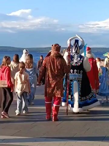 A group of people are walking along a promenade by a body of water under a cloudy sky. In the foreground, two young girls in casual clothing are walking away from the camera. Behind them, a person in a richly patterned brown coat and red boots is walking away from the camera. To their right, a person in an elaborate blue and white costume with a tall headdress and staff is walking away from the camera. To the right of that, a person in a red Santa Claus-like costume with a white trim is walking away from the camera, holding a staff. There are other figures in similar costumes, some appearing to be Santa Claus figures, walking in the same direction, as well as other people in casual clothing. A young girl in a blue dress is visible on the far right. The environment suggests a festive outdoor event.