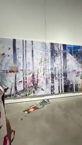 The video shows a large painting in an art gallery. The painting is mostly white, with what appears to be an abstract rendering of a forest, with streaks of other colors like blue and pink. On the floor in front of the painting is what looks like a smashed or melted object with wires or cords extending from it. A thin cable is hanging from the ceiling. The gallery has white walls and a polished concrete floor. Two people are seen in the video. One is a woman standing in the foreground, wearing a jacket and carrying a purse. The other is a person wearing dark clothes walking away from the camera near the wall. The room is well lit.