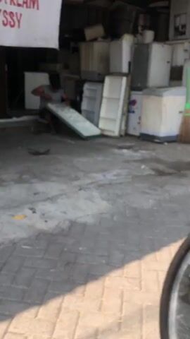 The video shows an outdoor scene with a person interacting with what appears to be discarded appliances. The person, wearing a grey t-shirt, is bent over and appears to be moving or examining a large, flat, reflective object, possibly a mirror or a piece of glass, that is propped against other items. Behind the person and to the right, a collection of refrigerators and other large appliances are stacked and leaning against each other. The ground is a mixture of concrete and what looks like salvaged materials. To the far right, a broom is visible. The background is cluttered with more appliances and structures, suggesting a storage or salvage yard. A portion of a white banner with red text is visible in the upper left corner. The lighting suggests it is daytime.