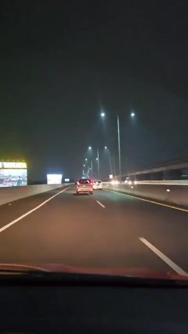 The video shows a view from inside a car driving on a well-lit highway at night.  Several cars are visible ahead in the same lane, and a concrete barrier runs along the right side of the road. There are streetlights on either side of the highway, and a large billboard is visible to the left. The windshield of the car is also visible.