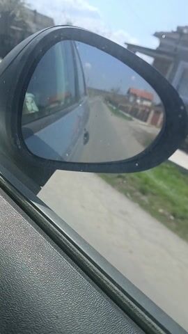 The video shows a car side mirror while driving down a narrow asphalt road in a rural or suburban area. The reflection in the mirror shows the back of the car and the road behind it, with a few houses and trees visible on either side. The sky is blue with some scattered clouds. The car is moving, and the scenery is passing by in the mirror's reflection. The road appears to be in a relatively quiet residential area.