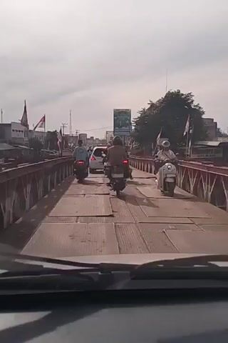 The video shows a view from a vehicle driving across a bridge. Multiple motorcycles and a car are visible on the bridge, traveling in the same direction. The bridge appears to be made of metal panels and has red railings on either side. Buildings and trees are visible on the sides of the bridge, and flags are flying. The sky is overcast.