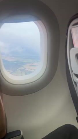 The video shows an interior view from an airplane window. A person wearing glasses is partially visible on the left side of the frame, their hand resting near their chin. Through the window, a landscape of fields, some green and some dry, and scattered buildings is visible below. The sky appears overcast or hazy. The right side of the frame shows a portion of the airplane's interior, including a black and silver object.