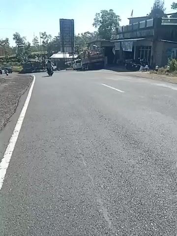 A truck has crashed into a building on the side of a road. The truck is tilted at an angle and appears to have sustained damage. A white van is parked behind the truck. A billboard stands behind the truck and van. Several motorcycles are on the road. A few cars are also visible in the distance. The weather is clear and sunny.