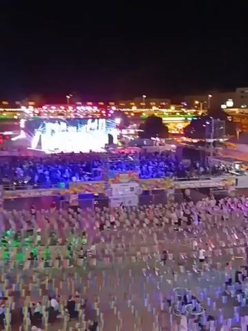 The video shows a large outdoor venue at night, likely for a concert or event. A crowd of people, many seated at tables, are watching a performance on a large stage lit with bright lights. There are several buildings surrounding the venue with lights and signage, suggesting it is a commercial area. The video is taken from a high vantage point, possibly a building overlooking the scene.