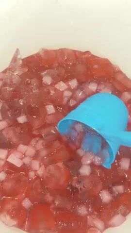 A close-up, overhead shot shows a blue plastic scoop stirring a mixture of red liquid, ice cubes, and small pink cubes in a white bucket. The pink cubes appear to be some type of soft candy or jelly. Tiny black seeds are dispersed throughout the mixture. The liquid is being stirred vigorously, creating ripples and splashes.