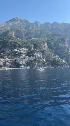 The video pans across a body of water, revealing a coastal town built into the side of a steep, verdant mountain. The town is characterized by numerous white buildings with colorful accents, densely packed together and cascading down the hillside. Several sailboats are visible anchored in the water near the shore. The sky is clear and blue, and the sun is shining, casting a bright light on the scene. The water is a deep blue, with small waves rippling across its surface. The overall impression is one of a picturesque Mediterranean landscape.