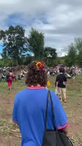 The video shows a dirt field filled with parked scooters and some cars. Several people are walking around the field, mostly dressed in casual summer clothing. The sky is blue with scattered clouds, and there are trees surrounding the open area. The ground is muddy and shows tire tracks from the vehicles.