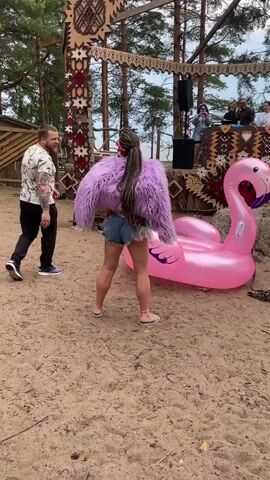 A woman wearing a fluffy purple jacket and denim shorts with her hair in a long braid is dancing near a large inflatable pink flamingo. Behind her, a man in a white patterned shirt and black pants is looking at her and smiling. In the background, there is a stage decorated with intricate patterns, with DJs and speakers set up. Several other people are visible on the stage and in the audience. The scene appears to be an outdoor event, possibly a festival, with sand on the ground and trees in the distance. The woman is facing away from the camera, her back to the audience and the stage. The inflatable flamingo is positioned to her right. Another woman is visible to the right of the frame, also dancing and facing the stage. A third woman is in the foreground on the left, crouching and holding up a phone to take a picture. The lighting suggests it is daytime, with an overcast sky.