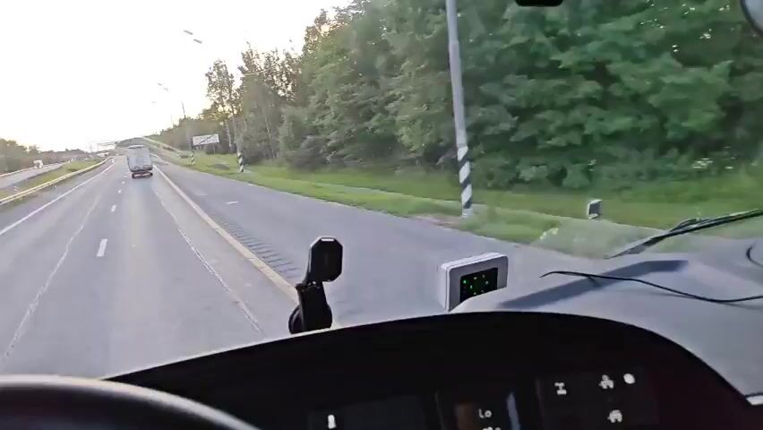 The video captures a first-person view from the driver's seat of a truck traveling on a highway. The road is paved and has multiple lanes, with a yellow line marking the shoulder and white lines dividing the lanes. The truck is in the right lane, and other trucks can be seen in the distance. The sky is bright, indicating daytime, with some trees lining the right side of the highway. A sign with Russian text is visible on the right side of the road. Inside the truck, a device with green lights is mounted on the dashboard. The steering wheel is partially visible in the foreground.