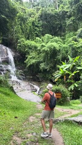 A person wearing a hat, an orange shirt, and khaki shorts stands facing a waterfall in a lush, green forest. They are holding a phone and appear to be taking a photo of the waterfall. A backpack is slung over their shoulders. A signpost is visible to the right of the person. The waterfall cascades down a rocky surface surrounded by dense vegetation. The ground is a mix of dirt path and green grass.