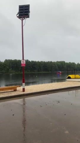The video pans across a waterfront area on a cloudy day. In the foreground, a wet, paved area with puddles suggests recent rain. A sandy embankment is visible, leading to a body of water. On the water, there are a few buoys and a brightly colored inflatable structure. Along the shore, there is a wooden bench and a dock with several yellow and blue pedal boats. Further down the dock, a large blue trash bin is situated near a red tent. People are seen walking along the path and sitting under the tent. The background features a dense line of trees. The video moves from left to right, showing a wider view of the area.