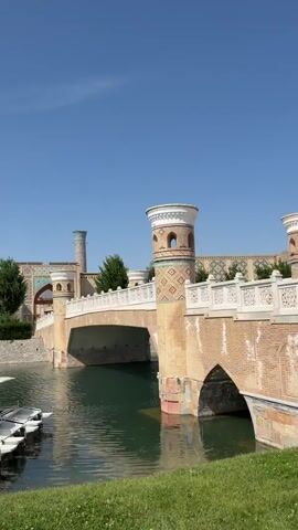 This image captures a serene daytime scene featuring an ornate bridge spanning a dark green canal or river. The bridge is constructed with brick-like material, featuring multiple arches and a light-colored railing. On the bridge, there are two prominent, decorative towers with intricate geometric patterns in brick and what appears to be blue and white tile work, topped with white and green elements.

In the background, behind the bridge, several buildings with distinct Central Asian architecture are visible. These buildings are adorned with colorful, intricate tile mosaics, and another tall, slender tower (resembling a minaret) rises on the left side. Green trees are interspersed throughout the landscape, both on the banks of the canal and behind the buildings.

On the left side of the canal, several small, white boats are moored, reflecting in the calm water. The foreground on the right features vibrant green grass, indicating a well-maintained area. The sky above is clear and blue, suggesting a bright, sunny day. The overall scene conveys a sense of cultural heritage and picturesque beauty, consistent with the location of Kuchtumgali, Uzbekistan. No people or visible text are present in the image.