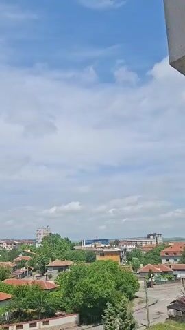 The video captures a panoramic daytime view of a town, likely from an elevated perspective such as a balcony or upper-story window. The setting is a mix of residential areas and more urban structures in the distance, consistent with a town like Galabovo, Bulgaria.

In the foreground and midground, numerous single to two-story houses with distinctive terracotta or red-tiled roofs are prominent. These homes are nestled among abundant lush green trees, suggesting a verdant environment. Further in the background, taller, multi-story apartment buildings and other urban structures punctuate the skyline. A road with vehicles can be briefly seen in the lower right portion of the frame.

The sky is mostly clear blue with scattered white, cumulus clouds, indicating fair weather and a bright, sunny day. Throughout the video, several small birds are visible flying high in the sky.

The primary activity is a slow, smooth horizontal pan of the camera from right to left, revealing more of the townscape.

A notable audio detail is a loud, distinct emergency siren that is heard throughout the first half of the video, peaking in intensity and then gradually fading. No discernible text is visible on any buildings or objects in the video.