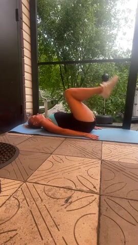 A woman is performing a bicycle crunch exercise on a blue yoga mat outdoors. She is lying on her back with her legs raised and bent at the knees. Her hands are behind her head. She is wearing a turquoise and black athletic top and black shorts. The mat is on a tiled patio. In the background, there is a wall, a doorway, trees, and a punching bag. The video shows the woman moving her legs in a pedaling motion, alternating bringing her knees towards her chest and extending her legs.