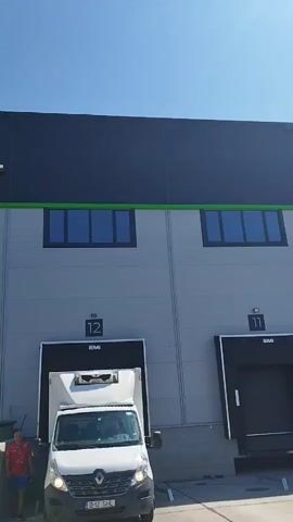 The video shows the exterior of a modern industrial building with a dark blue roof and light grey walls. The building has several loading dock bays, some open and some closed. A white refrigerated truck is parked at one of the loading docks, with a person standing beside it. Another person is visible near another loading dock. Two cars are parked in front of the building, and a small tree is planted near one of the entrances. The sky is clear and blue. The building has signs that say "Banca pentru Alimente Bucuresti" and "Homplex". The video pans from left to right, showing different sections of the building's facade.