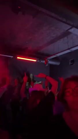 The video shows a dark, crowded nightclub with people dancing and raising their hands in the air. The lighting shifts from darkness to flashing red and blue lights, with haze filling the room. The crowd is mostly silhouetted against the light. Some individuals can be seen more clearly, including a person with glasses in the foreground, and others wearing white hats. The environment has exposed ceiling elements, suggesting an industrial or modern style.