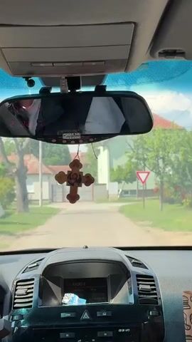 A view from inside a car looking out the front windshield. The car is stopped on a narrow, paved road in a residential neighborhood. The dashboard is visible, featuring a radio and ventilation controls. A wooden cross with an image is hanging from the rearview mirror. The mirror also reflects the car's interior, showing clothing. Outside, there are houses with gated driveways, trees, and a yield sign. The weather appears sunny.