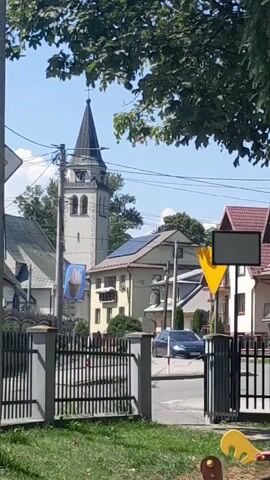 This is a slightly shaky, short video clip of an outdoor scene, likely a village or small town, on a sunny day. The main focus is a tall, light-colored church tower with a dark spire and a cross at its apex. The tower has arched windows. To the left of the tower, a portion of another building, possibly part of the church, is visible with a steeply pitched roof. A banner hangs from a cable in front of the church tower, featuring an image of an ice cream cone.

In front of the church buildings, there is a grey metal fence with vertical bars and wider grey posts. Beyond the fence, a street is visible with some houses on the right side. One house has a red tiled roof and a blank, square signpost in front of it. Another house has solar panels installed on its roof. There are utility poles and wires crisscrossing the scene. Trees with green foliage frame the top of the video, and some grass and possibly children's toys are visible in the foreground at the bottom. The sky is blue with some scattered white clouds.