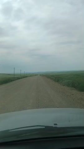 The video shows a point-of-view shot from inside a vehicle driving down a gravel road. The road stretches into the distance, flanked by green fields on either side. Utility poles are visible along the left side of the road. The sky is overcast with a cloudy sky. The dashboard and windshield wipers of the vehicle are visible at the bottom of the frame. The video appears to be filmed during the daytime.