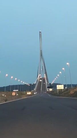 The video captures a view of a modern cable-stayed bridge from a roadway approaching it. The sky is a clear, pale blue, suggesting either dawn or dusk. Streetlights line the sides of the roadway, curving upwards as they approach the bridge, and are illuminated. Several cars are visible on the roadway, some driving towards the bridge and others in the distance on the bridge itself. The bridge has a tall, slender central pylon that leans dramatically, with cables extending downwards to support the roadway. The overall impression is one of impressive engineering and an approaching journey.