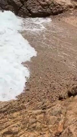 Here is a concise description of the video: 

The video shows crashing waves at the edge of a small beach area. The waves are white and foamy as they meet the rocky edge of the beach. The rocks are brown and rugged, and the sandy beach area is visible in the background. The video seems to be shot from an elevated position, possibly from the rocks themselves.
