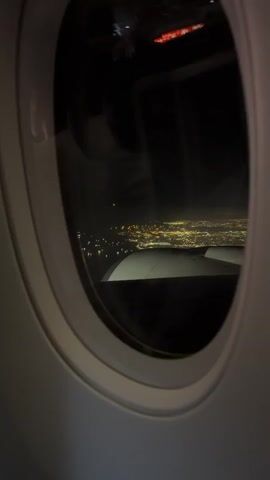 This media file captures a view from an airplane window at night, overlooking a sprawling cityscape illuminated by a multitude of lights. The oval-shaped window frames the scene, with the interior of the airplane's cabin faintly visible as reflections on the glass. The main subject is the distant city, a tapestry of yellow and white lights stretching across the horizon. A portion of the airplane's wing is visible in the foreground, its smooth, white surface contrasting with the darkness of the night sky. The activity is implied: an airplane in flight, likely cruising at altitude.

Notable details include the time of day, which is clearly nighttime, indicated by the darkness and the illuminated city. There are no obvious weather cues, but the clarity of the city lights suggests a clear night. Location cues are limited to the context provided: unknown, Turkey. The emotion conveyed is one of wonder and awe, a common feeling when observing a city from above at night, evoking a sense of scale and human presence.

There is visible text on a red light fixture at the top of the airplane cabin, visible in the reflection on the window. However, the text is too blurry to be read clearly.