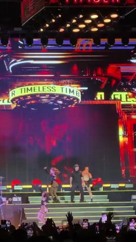 The video shows a live concert performance on a brightly lit stage. A male performer in a black shirt and hat stands center stage, interacting with several female dancers dressed in revealing outfits. Behind them, a large LED screen displays visuals, including a close-up of a man's face and other abstract imagery. The stage is decorated with large, circular light fixtures and a banner that reads "TIMELESS TIMBER." In the foreground, an audience is visible, with many people holding up their phones to record the event. The overall atmosphere is energetic and visually dynamic, typical of a large-scale concert.