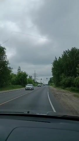 The video shows a view from inside a car driving on a two-lane road. The sky is cloudy. On the left side of the road, there are trees and a power line tower. On the right side, there are more trees. Several cars are visible on the road. There are also pedestrian crossing signs and traffic lights. The car is moving forward.