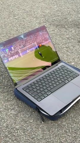 The video shows a group of young adults watching a soccer match on a laptop outside. The laptop is placed on a blue case on the ground. The camera zooms in on the screen displaying a soccer game, with brands like Pepsi and Mastercard visible. The group is seated on a bench near a chain-link fence. Some of them are smoking, and there are bags and cans scattered around. The environment appears to be an outdoor recreational area. The group celebrates and cheers as a goal is scored in the game.