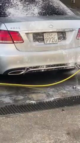 The video shows a silver Mercedes-Benz sedan being washed in an indoor car wash bay. The car is covered in soapy foam. A person wearing jeans and white rubber boots is using a yellow high-pressure hose to rinse the car, starting from the rear. A gray wall is visible in the background, and a ladder leans against the wall on the left. The car wash bay has a grated floor.