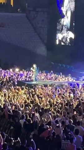The video shows a large crowd of people at a concert. A performer is on stage, singing into a microphone. The crowd is very enthusiastic, with many people holding up their phones to record the performance. The stage is lit up with bright lights, and there are large screens displaying images of the performer. The concert appears to be taking place in a stadium or arena, with tiered seating visible in the background. The atmosphere is energetic and exciting.