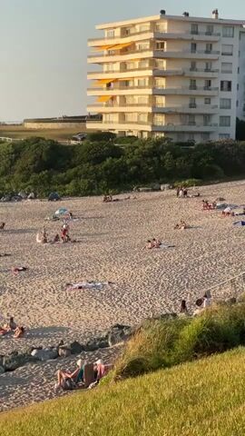 The video shows a sandy beach with people sunbathing and relaxing. In the background, there is a multi-story white building with balconies and a line of trees. There are also some small structures with thatched roofs on the beach. The lighting suggests it is late afternoon or early evening, with the sun casting long shadows. The overall atmosphere appears peaceful and leisurely.