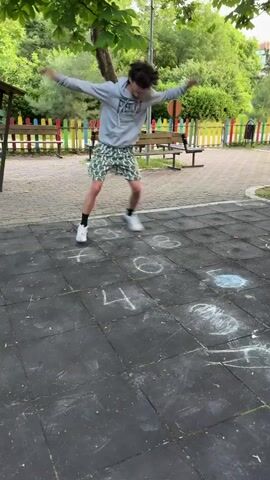 A young man with dark curly hair wearing a grey hoodie and patterned green shorts plays hopscotch outdoors. He is in mid-air, jumping from one square to another. The hopscotch grid is drawn on a dark rubberized surface with chalk. Behind him, a colorful playground fence with yellow, red, blue, and green posts is visible, along with a wooden bench and trees. Another person, partially visible, stands to the right. The ground is a textured dark gray, with chalk drawings of numbers and figures. The young man is in motion, with his arms outstretched and his body angled as he jumps. He is wearing white sneakers.