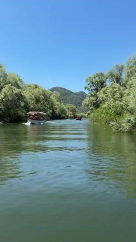 A video of a green river with boats on it, with trees and mountains on either side. There are three boats moving away from the camera. The boat closest to the camera is on the left side of the river. The boat is white with a brown canopy. There are people in the boat. The other two boats are smaller and further away. The sky is blue and clear. The water is calm and there are ripples from the boats. The trees on either side of the river are green and lush. The mountains in the background are green and have some rocky parts.