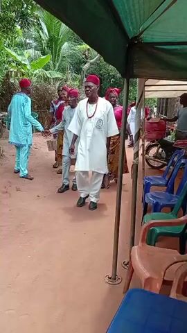 The video shows a group of people walking under a green canopy. Many of them are wearing traditional Nigerian clothing. There are plastic chairs arranged along one side of the walkway. There is a bicycle to the left of the frame. The ground is red.