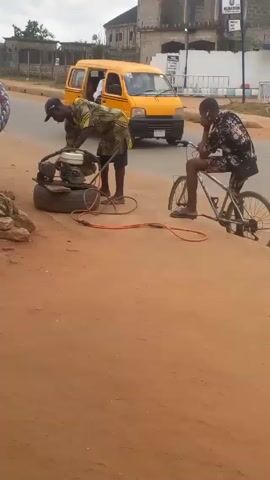 The video shows two men on a roadside in what appears to be a tropical or subtropical location. The road is paved and dry, with a reddish dirt shoulder. One man, wearing a patterned yellow and black shirt, dark shorts, and a baseball cap, is bending over an air compressor that is sitting on a tire. He appears to be starting or operating the compressor. The other man is seated on a bicycle with flat tires. He is wearing a black shirt with a floral pattern and appears to be looking at his phone. In the background, there is a yellow van partially visible and buildings across the street. The sky is overcast.