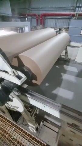 The video shows a close-up of a large industrial machine with two massive rolls of what appears to be brown paper or cardboard. The rolls are mounted horizontally and appear to be part of a larger system within a factory or industrial setting. The machine is primarily white and grey, with visible mechanical components. Red pipes run along the ceiling in the background. The overall environment looks like a warehouse or production facility. There are no people visible in the frame.