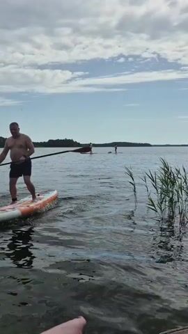 A man is paddleboarding on a lake. He is standing on a colorful paddleboard and holding a paddle. He is wearing black shorts. There are other people paddleboarding in the background. The sky is cloudy and there are trees on the horizon. The water is calm with some ripples. There is tall grass growing on the right side of the frame.