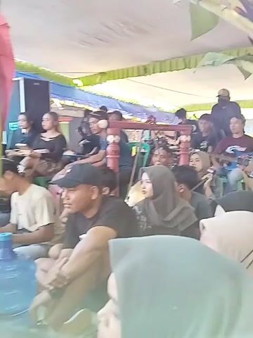 Here's a description of the video:

The video shows a group of people, mostly sitting, under a tent-like structure. Some individuals are wearing head coverings. A band appears to be performing; musical instruments are visible, including a guitar and percussion instruments.  The setting appears to be outside. Some people in the crowd seem to be watching the performance, while others are looking elsewhere or talking. The area around the tent has elements of greenery, including what seems to be a banana plant.